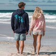 Man and women walking the beach. Man is wearing a navy hoodie with teal manta ray design on the back by  Studio Bilby. Women is wearing light pink hoodie with 'dive deep & rise above' mermaid design on the back by Studio Bilby.