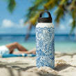 Stainless steel insulated water bottle with 'Blue Ocean' design by Studio Bilby. Bottle sitting on the sand of a tropical beach.  Background includes the sea, palm trees and people sunbathing.