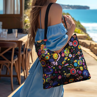 Women wearing a blue dress holding the playful pizzazz dark tote bag by Studio Bilby. Background of beachside cafe.